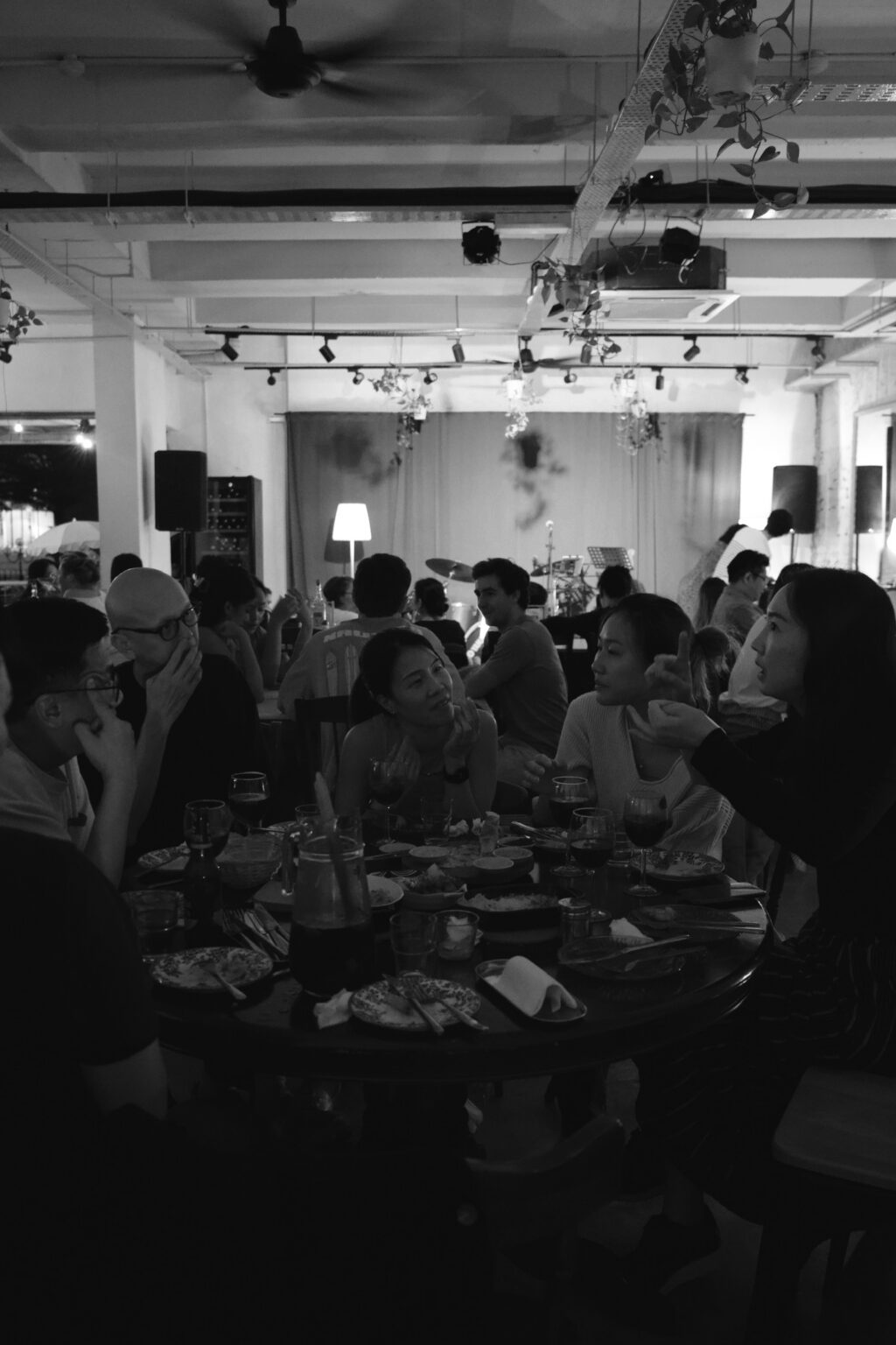 Black-and-white scene at Bartolo — friends chatting around a shared dinner table, stage in background